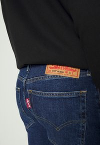 Jeans in denim blu scuro con vita alta. Presentano un'etichetta in pelle beige, una linguetta rossa del marchio e cuciture gialle sulle tasche.