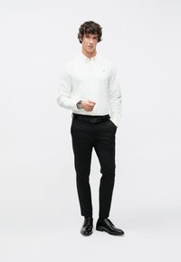 Weißes Hemd mit Knopfleiste und kleinem, gesticktem Logo, schwarze Hose und schwarze Anzugschuhe, mit schmaler Passform und glatter Stoffstruktur.