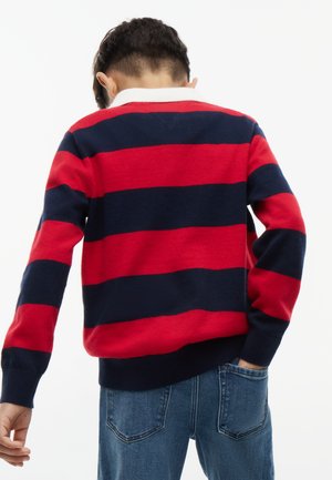 Barn iført rød og navyblåt stribet langærmet skjorte og blå jeans, stående med ryggen mod kameraet og den ene hånd i lommen.