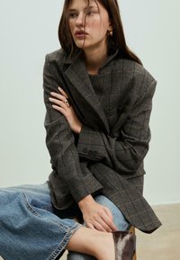 Blazer en chevrons marron à la coupe décontractée, avec une poche poitrine unique et des lignes à carreaux contrastantes, associé à un jean large bleu clair.