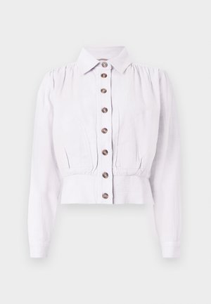 Blouse blanche à manches longues avec boutons marron à l'avant, taille ajustée, épaules légèrement bouffantes et col classique sur fond neutre.