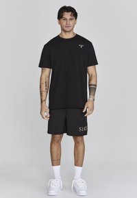 Black t-shirt, short sleeves, with logo; black shorts featuring "SIKSLK" in gold; white sneakers; plain background, standing male model.
