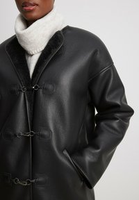 Cappotto in pelle nera con collo a imbuto e due chiusure a toggle. Il colletto interno è realizzato in un morbido tessuto grigio a contrasto.