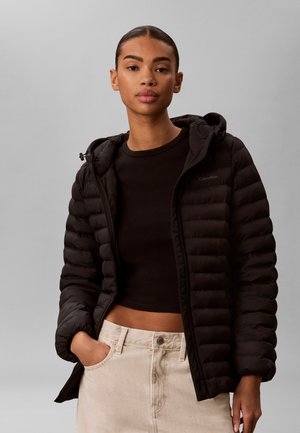 Mladá žena nosiaca čiernu prešívanú bundu cez čierny crop top a svetlo béžové nohavice, stojaca na jednolivom sivom pozadí.