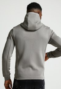 Felpa grigia con collo alto e cappuccio regolabile. Texture morbida con maniche attillate e dettagli nelle cuciture laterali. Vestibilità casual e rilassata.