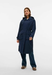 Marinfärgad vattentät jacka med huva, dragsnodd i midjan, två framfickor, knäppning och långa ärmar. Buren över lösa jeansbyxor.