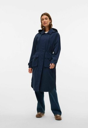 Veste imperméable marine avec capuche, taille à cordon, deux poches avant, fermeture à bouton et manches longues. Portée sur un pantalon en denim ample.