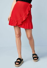 Jupe portefeuille rouge avec détails à volants, tissu texturé et lien ajustable à la taille. Associée à des sandales à plateforme noires.