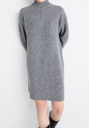 Szara sukienka z dzianiny prążkowanej sięgająca do kolan, z długimi rękawami i półzamkiem przy kołnierzu, noszona z czarnymi wysokimi butami sięgającymi do kolan.
