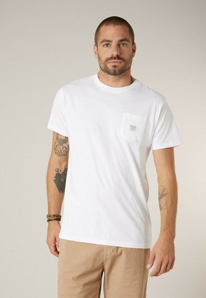Homme aux cheveux courts et à la barbe, portant un T-shirt blanc avec poche et un pantalon beige, montrant des tatouages sur les deux bras et des bracelets en perles.