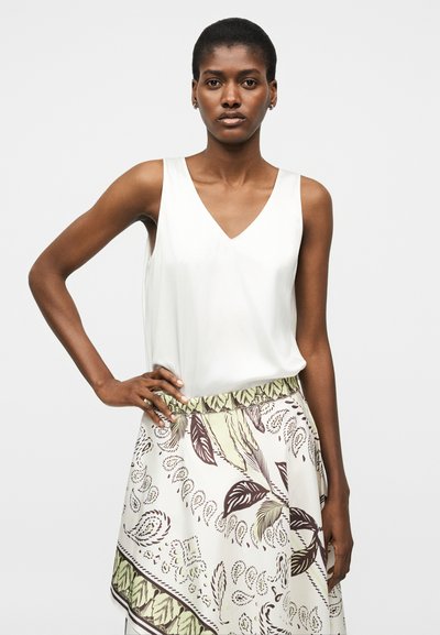 Γυναίκα με κοντά μαλλιά, που φοράει λευκό αμάνικο μπλουζάκι με V λαιμόκοψη και φούστα με σχέδιο καφέ και πράσινα φύλλα.