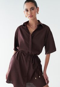 Brun kortærmet skjortekjole med knapper foran og bindebælte. Fremstillet i et tekstureret stof, har en krave og sideslidser.