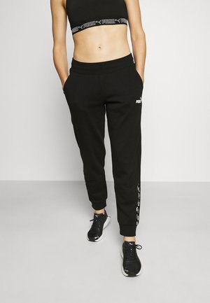 Femme portant un soutien-gorge de sport noir Puma, un pantalon de jogging noir Puma avec logo sur la cuisse et bande latérale, et des baskets noires Puma, debout les mains dans les poches.