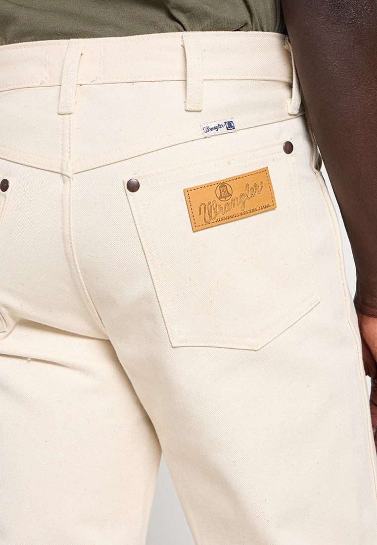Primer plano de una persona que lleva jeans Wrangler color marfil con un bolsillo trasero visible, parche de cuero de la marca y parte de una camisa verde oliva.