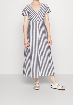 Kobieta nosząca szerokie, prążkowane kombinezon z krótkimi rękawami i dekoltem w serek, połączony z białymi sandałami z odkrytymi palcami i paskami.