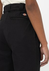 Pantalones de algodón negros con una cintura alta, bolsillo trasero con un logo de Dickies en rojo y amarillo, y un diseño de pierna recta.