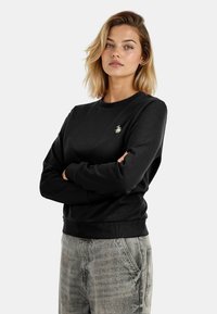 Black sweatshirt with ribbed cuffs and hem, featuring a small gold logo on the chest. Material appears soft, with a classic crew neck design.