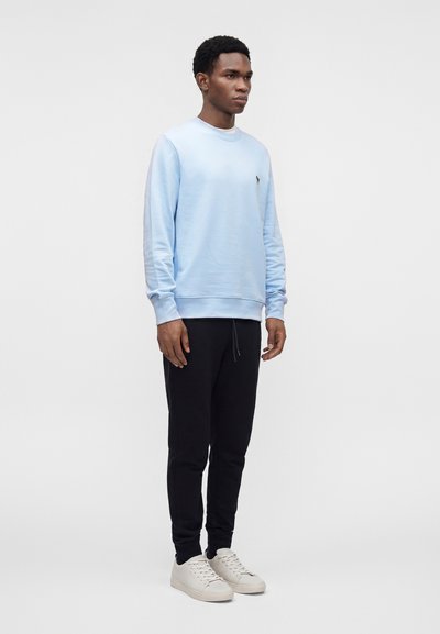 Homme debout portant un sweat-shirt bleu clair, un jogging noir et des baskets blanches sur un fond uni gris clair.