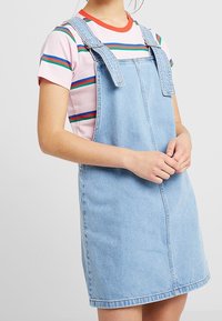 Denim hängselklänning i ljusblått med justerbara axelremmar, som har en framlomme och rak skärning, bärs över en randig T-shirt.