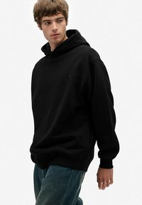 Sudadera negra con capucha, de manga larga, fabricada en un tejido suave, de corte relajado y con un pequeño logo en el pecho. Combinada con jeans de mezclilla azules.