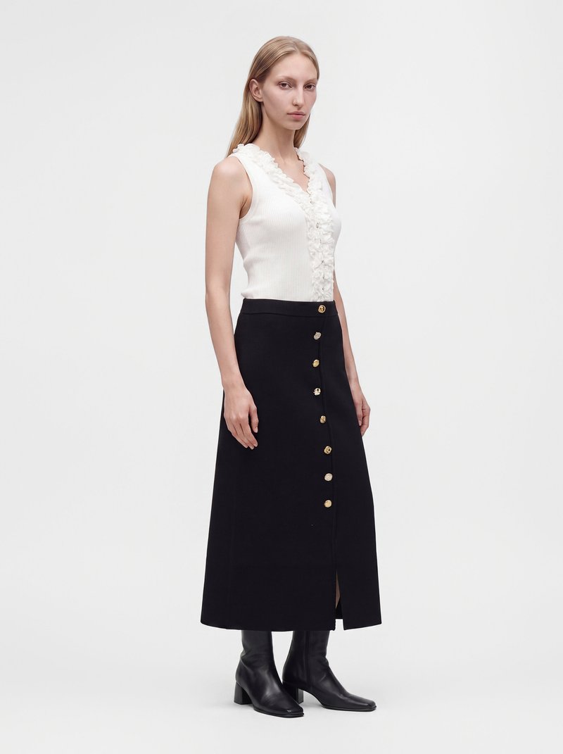 Woman wearing a white sleeveless top with ruffles, a black buttoned midi skirt, and black heeled ankle boots, standing on a plain background.