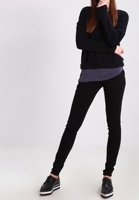 Svarta snäva byxor i kombination med en mörk strukturerad tröja. Modellen bär svarta skor med vita detaljer. Neutral bakgrund.