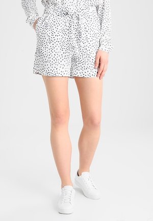 Femme portant un short blanc à motifs et des baskets blanches, debout avec une main dans la poche sur un fond uni.