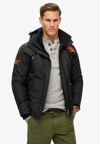 Czarna kurtka puchowa z wysokim kołnierzem, zapinanymi na zamek kieszeniami oraz pomarańczowym akcentem logo. Nosi ją na szarej koszulce henley. Widoczne oliwkowe spodnie.