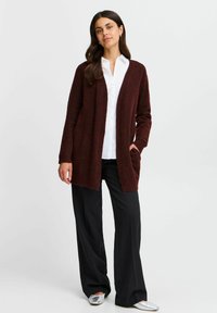 Cardigan bordeaux in morbido knit testurizzato con apertura frontale, maniche lunghe e tasche laterali, abbinato a una camicia bianca e pantaloni neri.