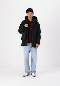 Sort isoleret jakke med hætte, båret over en bordeaux skjorte, parret med lyseblå jeans og sort-hvide sneakers. Lille sort taske fastgjort.