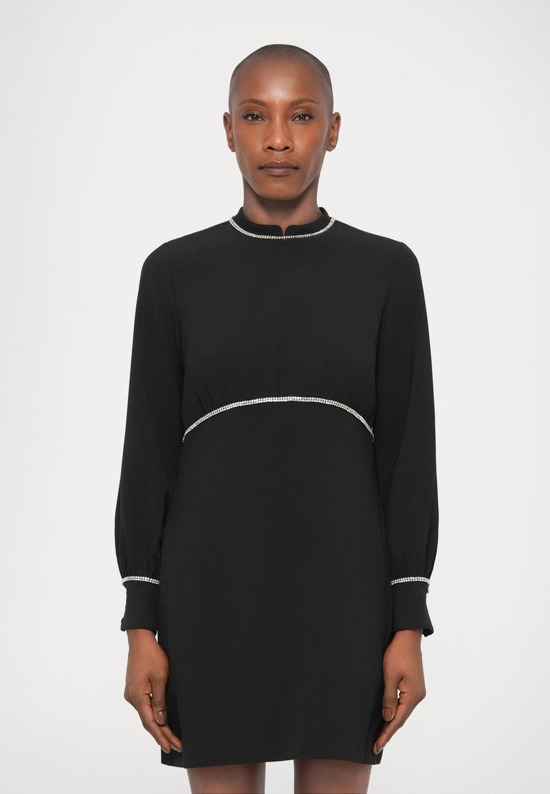 Vestido negro de manga larga y cuello alto, con una cintura ajustada y adornos plateados a lo largo del escote y la cintura.