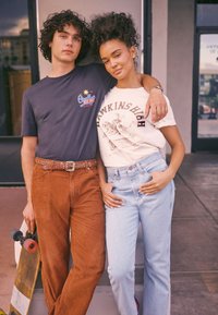 Poros asmenų poza lauke. Vienas dėvi tamsią marškinėlę su grafiniu raštu ir rudus korduroinius kelnes, kitas baltą marškinėlę su grafiniu raštu ir šviesios spalvos džinsus. Šalia guli riedlentė.