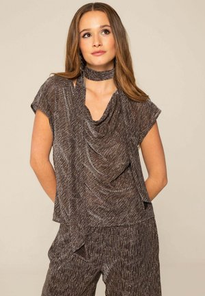 Mujer con una blusa marrón texturizada de escote drapeado y pantalones a juego de cintura alta, de pie con las manos detrás de la espalda sobre un fondo liso.