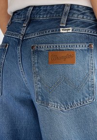 Pohľad zblízka na zadný vrecko modrých džín Wrangler so svetlohnedou koženou nášivkou s logom a výrazným šitím.