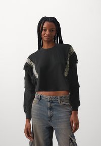 Felpa nera cropped con accenti a pattern e frange sulle spalle. Maniche lunghe con vestibilità morbida. Indossata con jeans a vita alta e scoloriti.