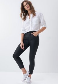 Jeans negros ajustados de cintura alta, combinados con una camisa blanca de botones. El modelo lleva zapatillas blancas, creando un atuendo casual.