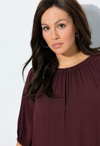 Bordeaux blouse met een textuurafwerking, losse pasvorm en gathered halslijn. Beschikt over mouwen tot de elleboog en een gladde, vloeiende uitstraling.