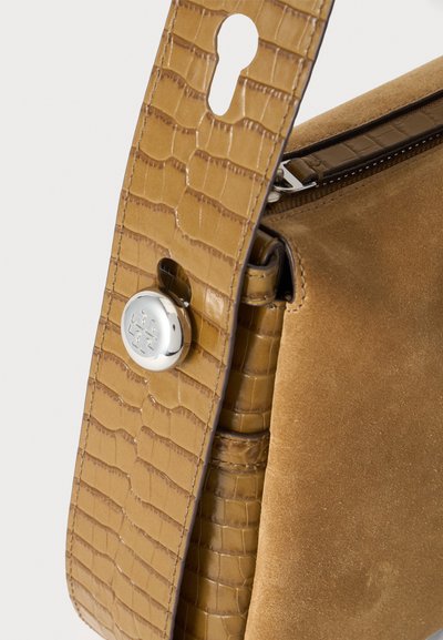 Sac à main en cuir beige et daim avec une sangle texturée présentant une boucle circulaire argentée et un détail de fermeture éclair sur le côté.