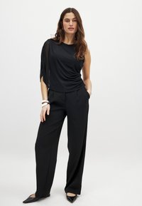 Sort top med én skulder, der draperer blødt, med en struktureret ærmedetalje. Sammen med højtaljede sorte bukser og spidse sorte flade sko.