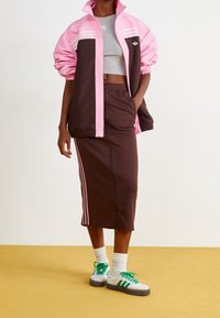 Brun idrottsbyxa med rosa ränder, i kombination med en rosa och brun jacka med ett subtilt mönster. Vita sneakers med gröna snören.