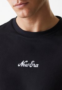 Black T-shirt in cotton fabric featuring a round neck and a white "New Era" logo printed on the chest. Minimalistic and smooth texture.