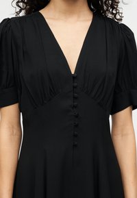 Μαύρο φόρεμα με βαθύ V λαιμόκοψη, κοντά φουσκωτά μανίκια και μπροστινό κούμπωμα με κουμπιά. Το ύφασμα έχει λείο, ελαφρώς υφή φινίρισμα.