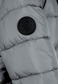 Siva puffer jakna s rukavima s teksturiranom crnom okruglom oznakom s inicijalima "NMT" i brojem "97." Glatka, sjajna tkanina, debljina s ukrasnim šavovima.