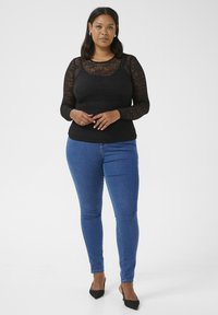 Camiseta negra de manga larga con patrón texturizado, combinada con jeans ajustados azules y tacones de punta negra. Diseño simple, forma ceñida.