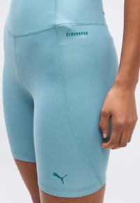 Lähedalt vaadatuna inimene, kes kannab helerohelisi Puma Cloudspun sportlühikesi pükse, kus on nähtavad õmblused ja logod, käsi rahulikult kõrval.