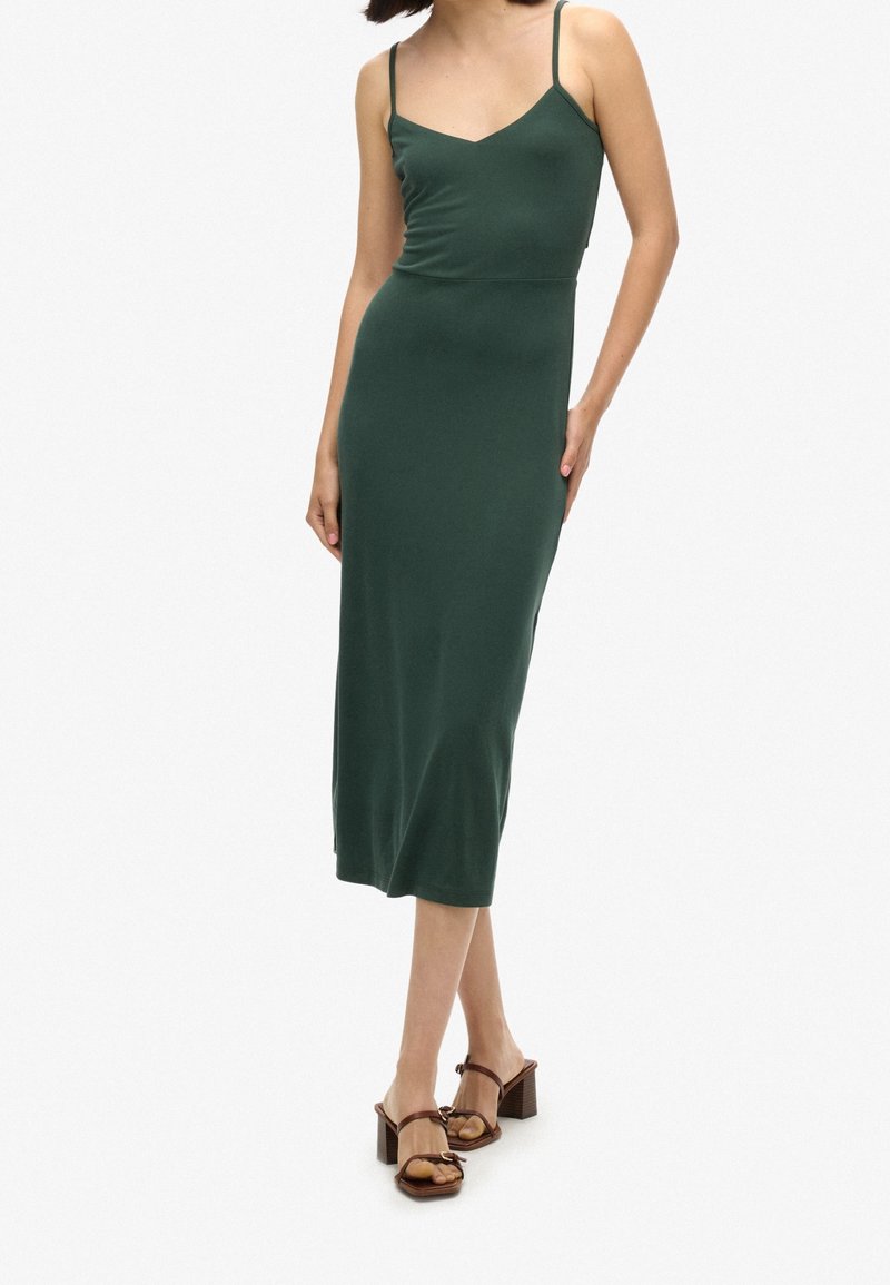 Femme portant une robe midi vert foncé sans manches avec de fines bretelles et des sandales à talons marron, debout devant un fond clair uni.