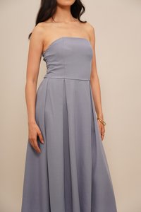 Ženska v brezramni svetlo vijolični obleki s plisejsko krilo in zlatimi zapestnicami na desni roki pred bež ozadjem.