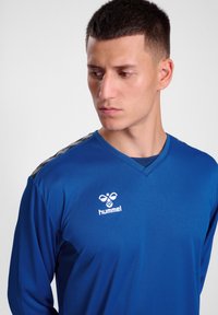 Blaues Langarm-Sportshirt mit V-Ausschnitt, das ein weißes Logo auf der Brust und graue Akzente an den Schultern aufweist. Glatte Textur.