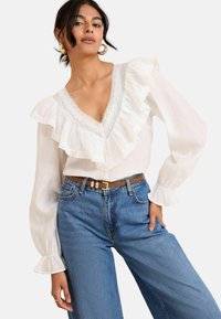 Blusa bianca con colletto e polsini arricciati, rifinita con pizzo. Abbinata a jeans in denim blu a vita alta e una cintura color sabbia.