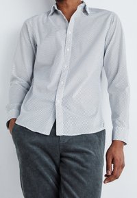 Camicia bianca con motivo a piccoli pois neri, maniche lunghe e colletto appuntito; abbinata a pantaloni in velluto a coste grigi testurizzati.